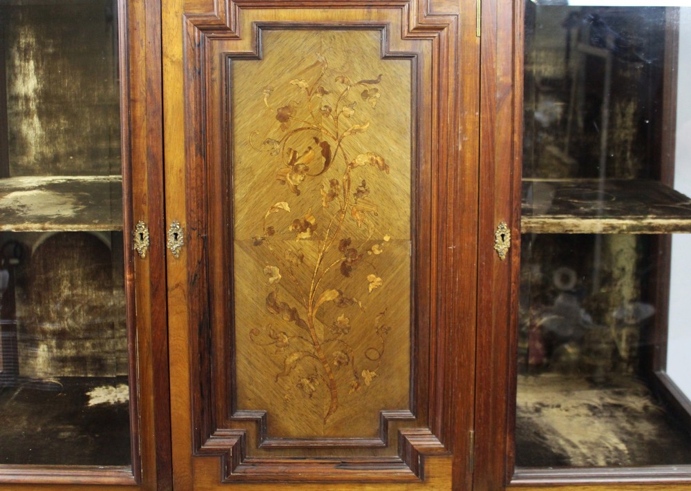 Late Victorian rosewood display cabinet featuring a central inlaid floral door, two glass side doors, a brass gallery rail top, and a lower tier shelf on turned legs. View IV