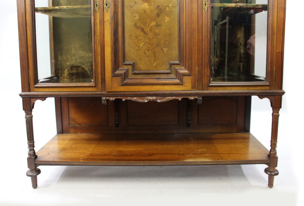 Late Victorian rosewood display cabinet featuring a central inlaid floral door, two glass side doors, a brass gallery rail top, and a lower tier shelf on turned legs. View V
