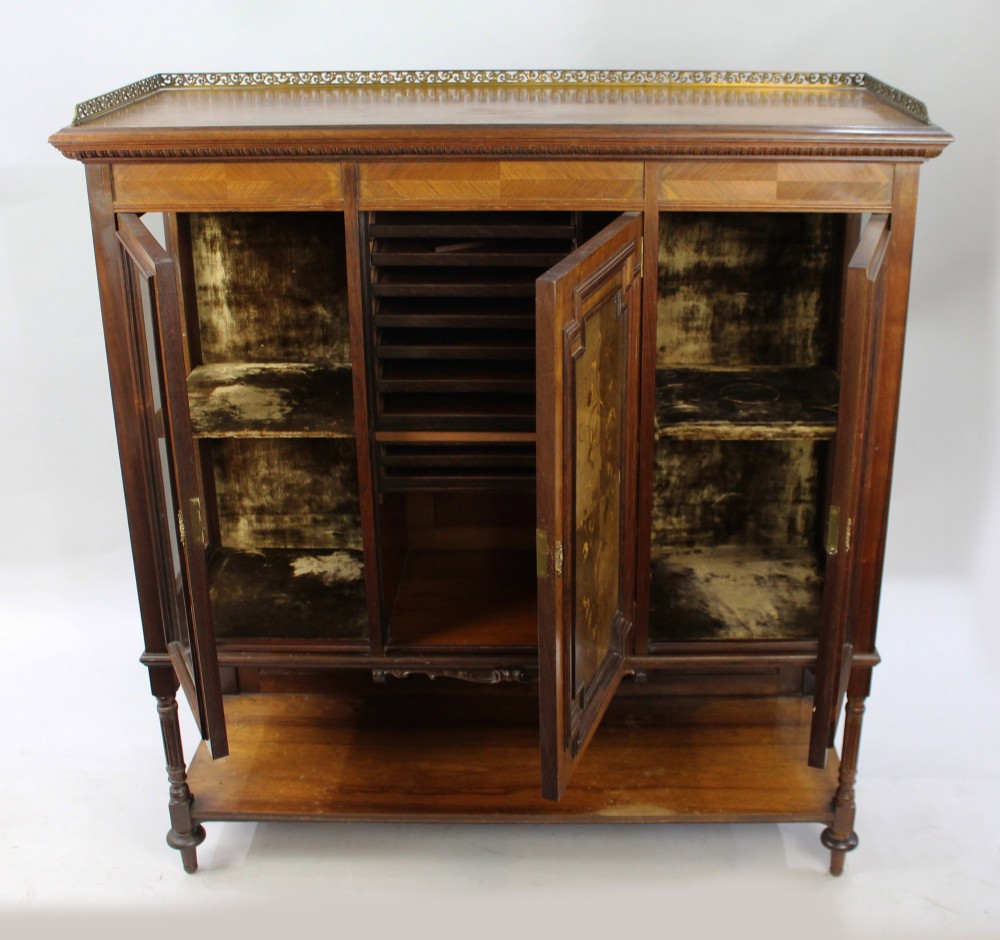 Late Victorian rosewood display cabinet featuring a central inlaid floral door, two glass side doors, a brass gallery rail top, and a lower tier shelf on turned legs. View VI