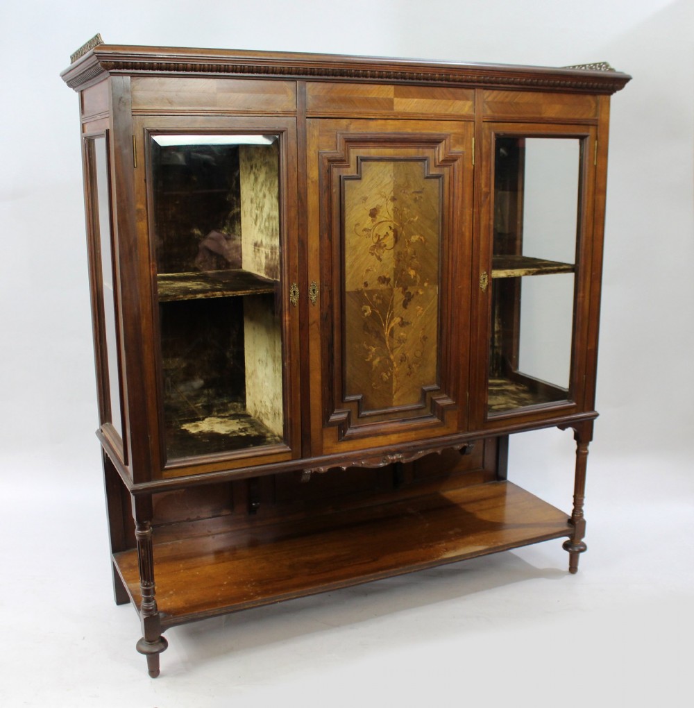 Late Victorian rosewood display cabinet featuring a central inlaid floral door, two glass side doors, a brass gallery rail top, and a lower tier shelf on turned legs. View VII