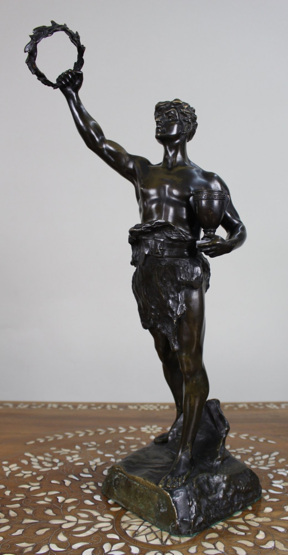 A dark brown patinated bronze figure of a classical male standing on a rocky base, holding a chalice in one hand and raising a laurel wreath aloft in the other.