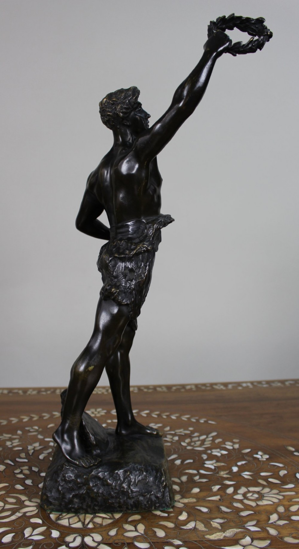A dark brown patinated bronze figure of a classical male standing on a rocky base, holding a chalice in one hand and raising a laurel wreath aloft in the other. View III
