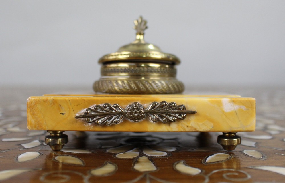 A rectangular yellow Siena marble desk inkstand featuring a central patterned ormolu inkpot with a finial lid, a front brass leaf applique, and small turned metal feet.