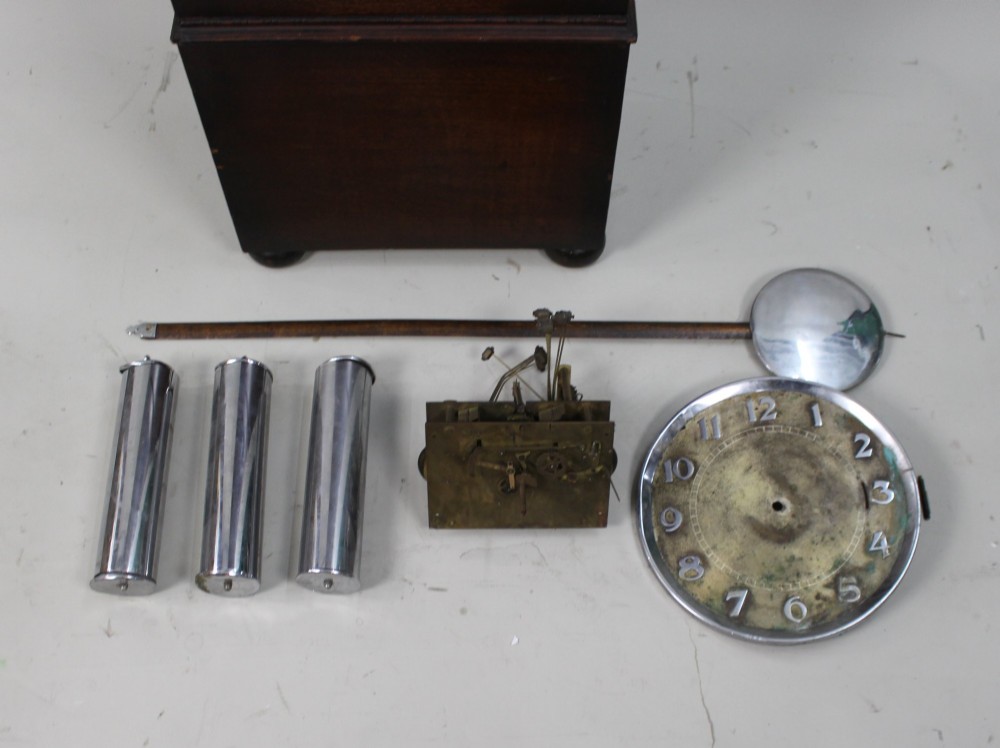 An Art Deco dark oak longcase clock case featuring a domed hood and glazed door alongside an unmounted brass movement, pendulum, chrome dial, and three cylindrical weights. View II