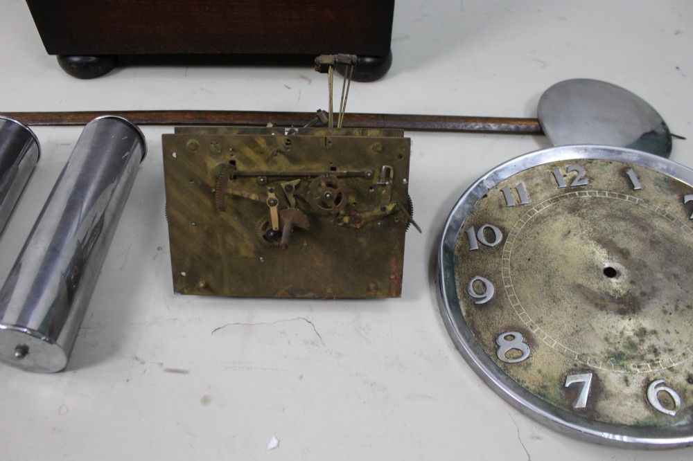 An Art Deco dark oak longcase clock case featuring a domed hood and glazed door alongside an unmounted brass movement, pendulum, chrome dial, and three cylindrical weights. View III