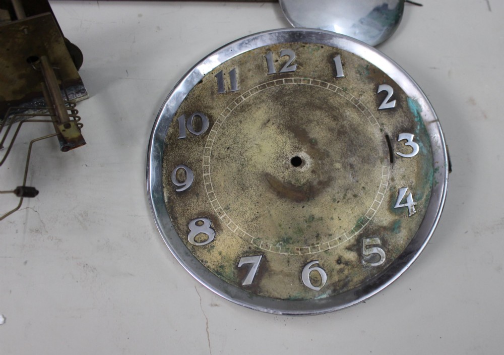 An Art Deco dark oak longcase clock case featuring a domed hood and glazed door alongside an unmounted brass movement, pendulum, chrome dial, and three cylindrical weights. View V