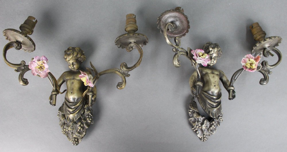A pair of cast metal wall sconces featuring sculpted cherubs holding twin scroll arms decorated with ceramic floral motifs and electrical light sockets.