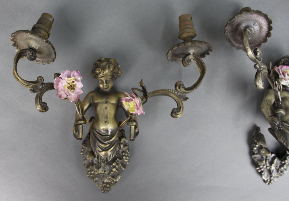 A pair of cast metal wall sconces featuring sculpted cherubs holding twin scroll arms decorated with ceramic floral motifs and electrical light sockets. View II