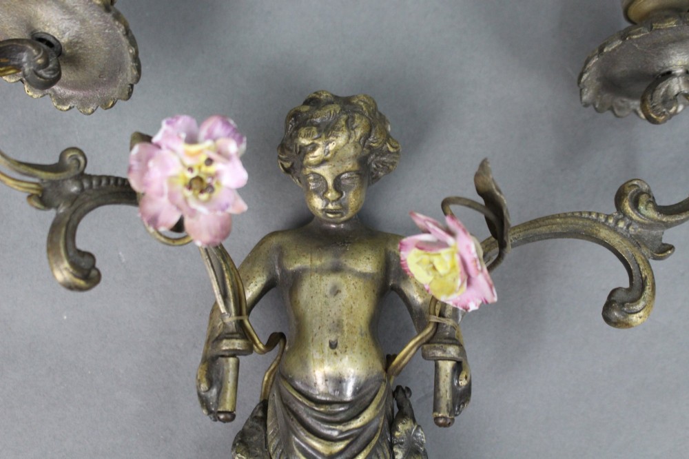 A pair of cast metal wall sconces featuring sculpted cherubs holding twin scroll arms decorated with ceramic floral motifs and electrical light sockets. View III