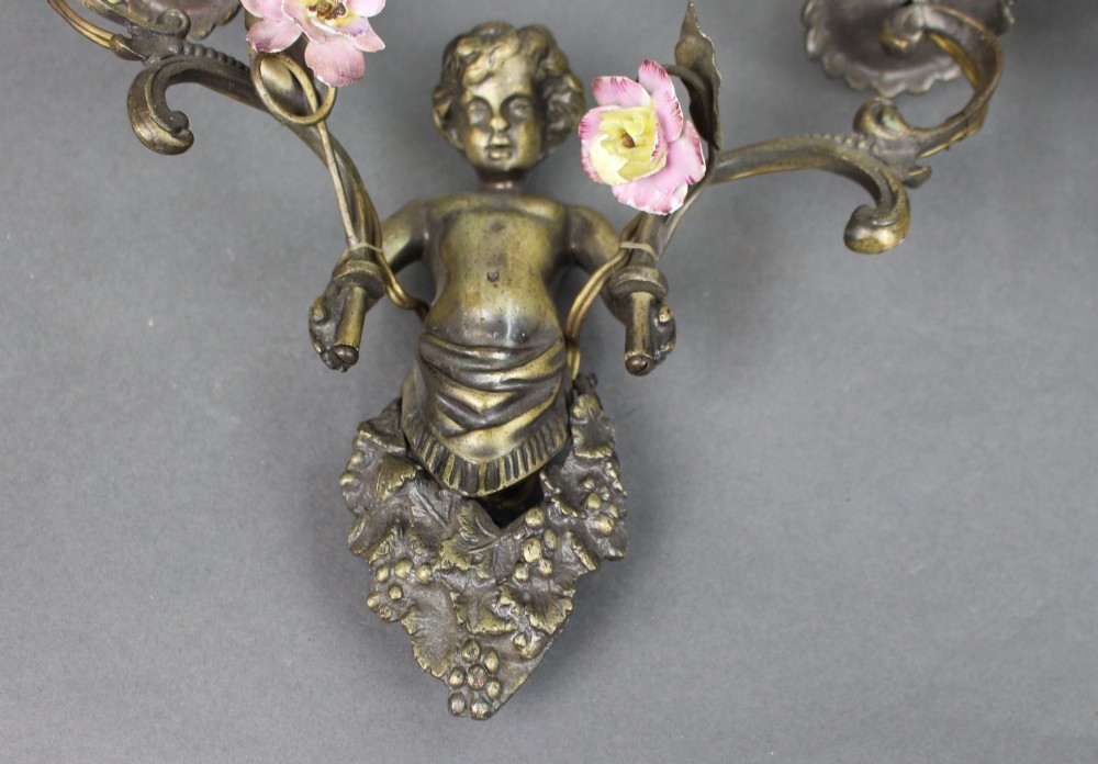 A pair of cast metal wall sconces featuring sculpted cherubs holding twin scroll arms decorated with ceramic floral motifs and electrical light sockets. View IV