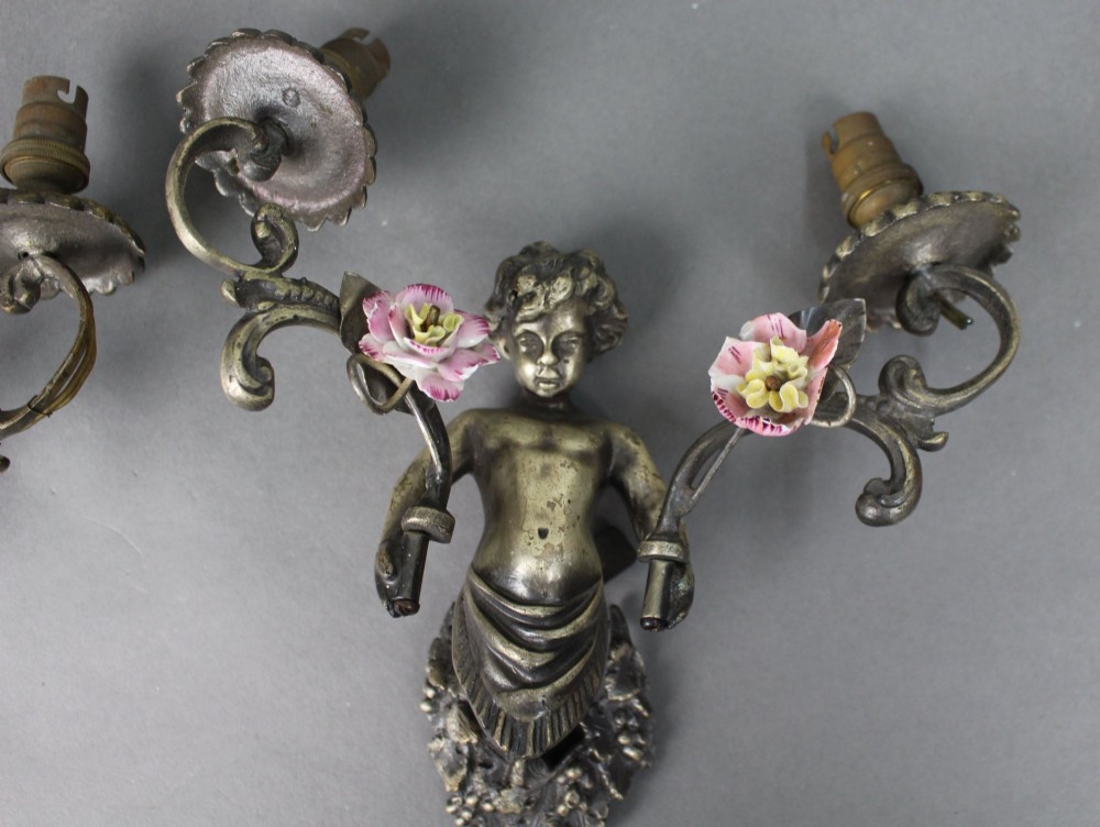 A pair of cast metal wall sconces featuring sculpted cherubs holding twin scroll arms decorated with ceramic floral motifs and electrical light sockets. View V