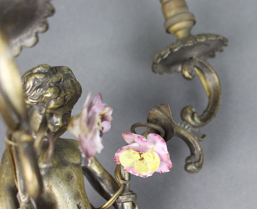 A pair of cast metal wall sconces featuring sculpted cherubs holding twin scroll arms decorated with ceramic floral motifs and electrical light sockets. View VIII