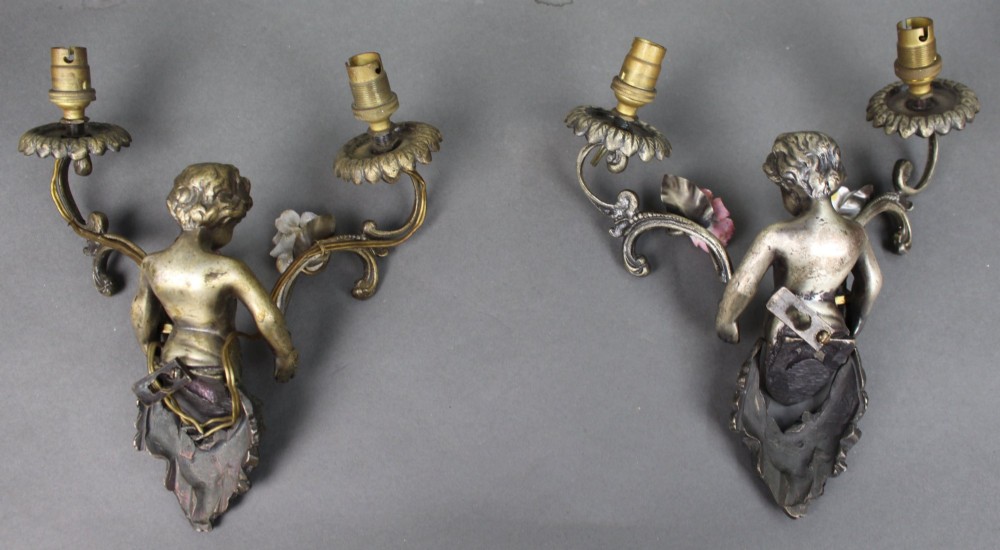 A pair of cast metal wall sconces featuring sculpted cherubs holding twin scroll arms decorated with ceramic floral motifs and electrical light sockets. View IX