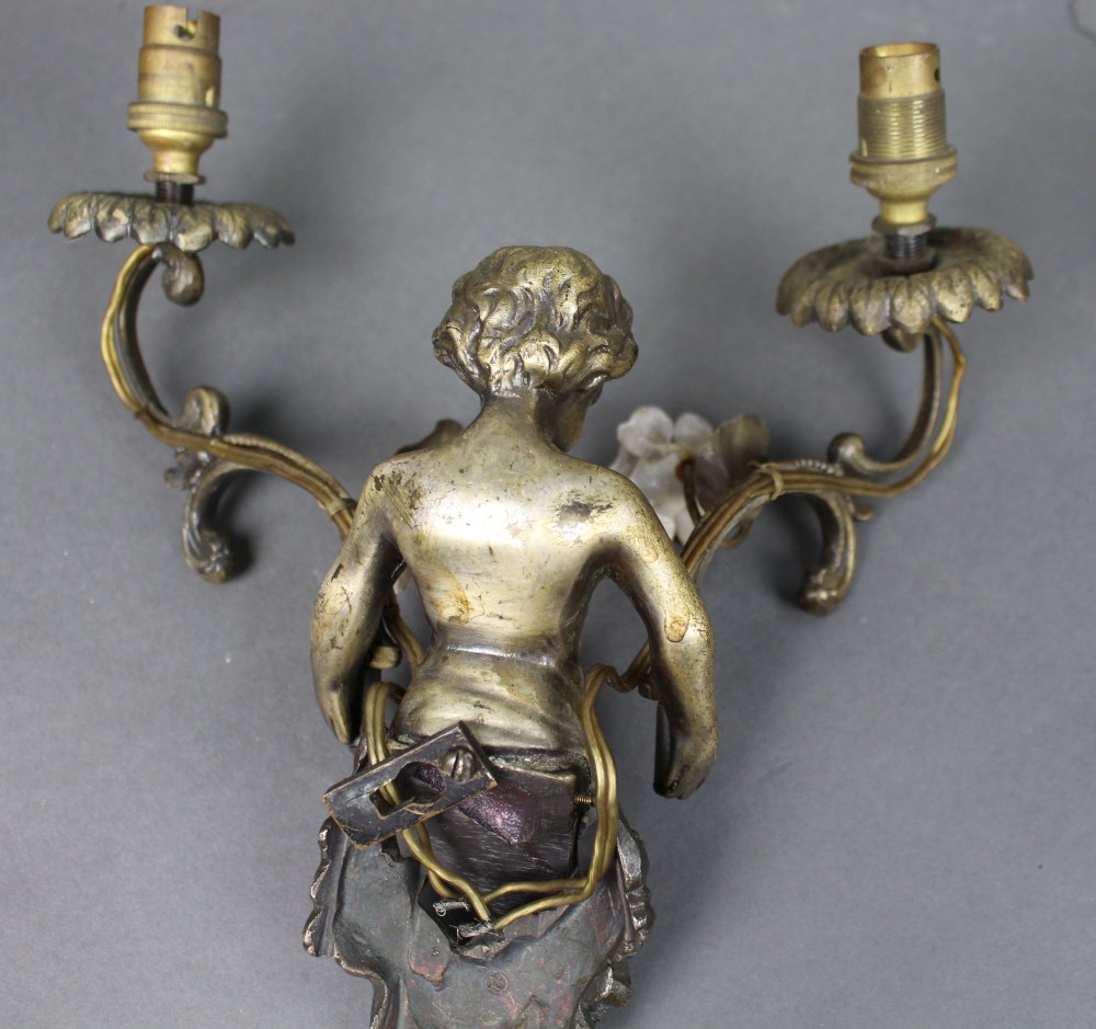 A pair of cast metal wall sconces featuring sculpted cherubs holding twin scroll arms decorated with ceramic floral motifs and electrical light sockets. View X