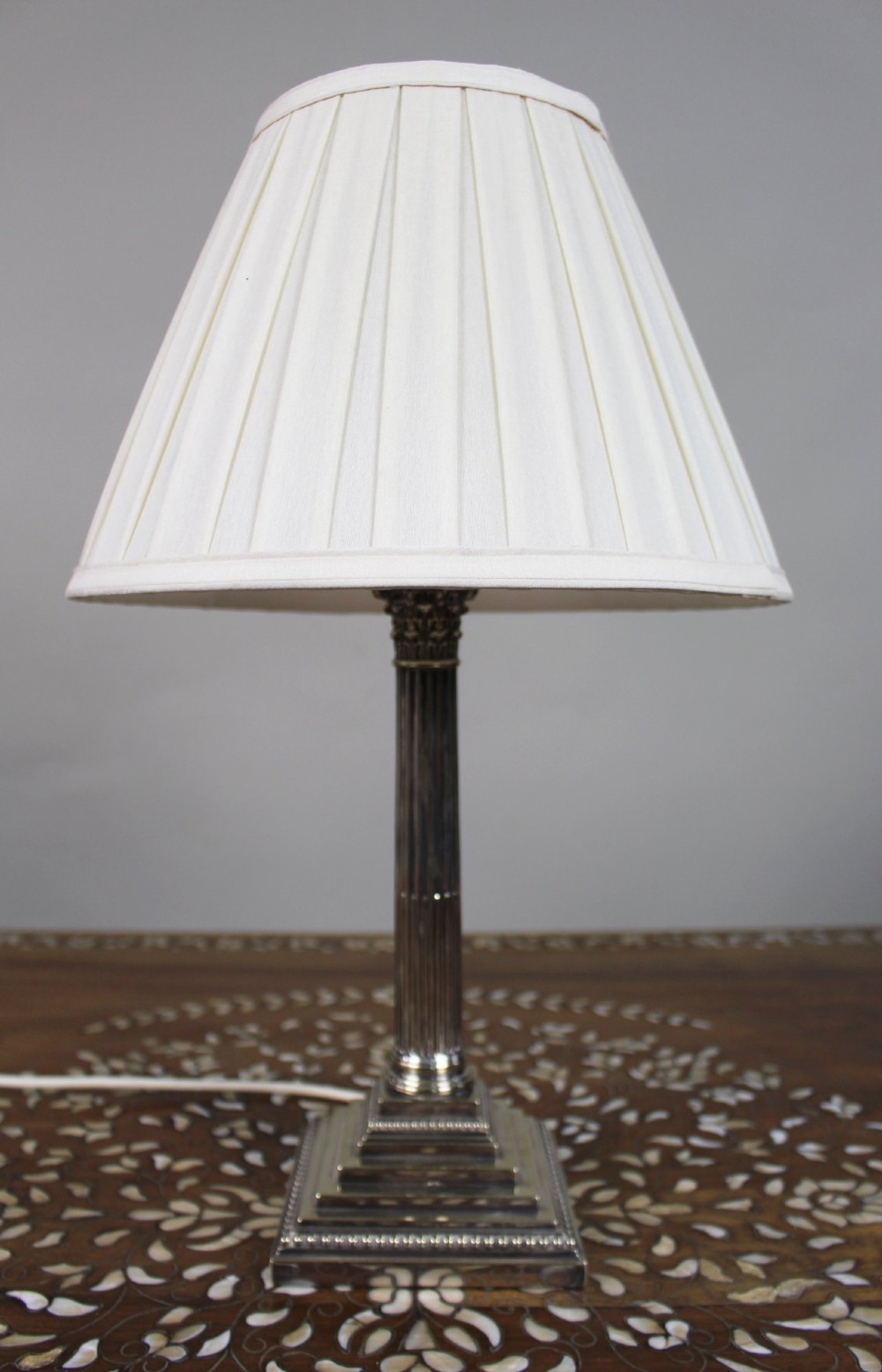 A silver-plated table lamp featuring a fluted Corinthian column shaft, a stepped square base, and a cream pleated fabric shade.