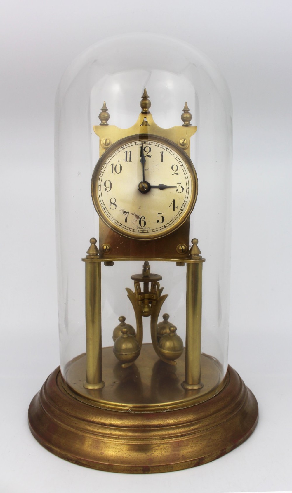 An antique German anniversary clock featuring a brass movement and four-ball rotary pendulum enclosed under a clear glass dome.