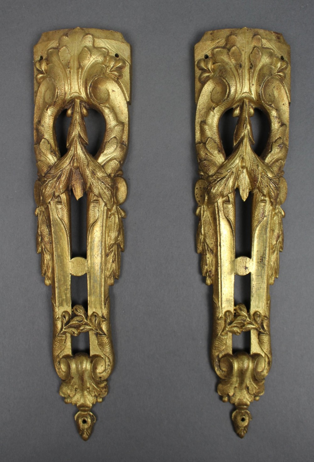 A pair of ornate gilt bronze furniture mounts featuring acanthus leaf scrollwork and symmetrical pierced design in the Louis XVI style.