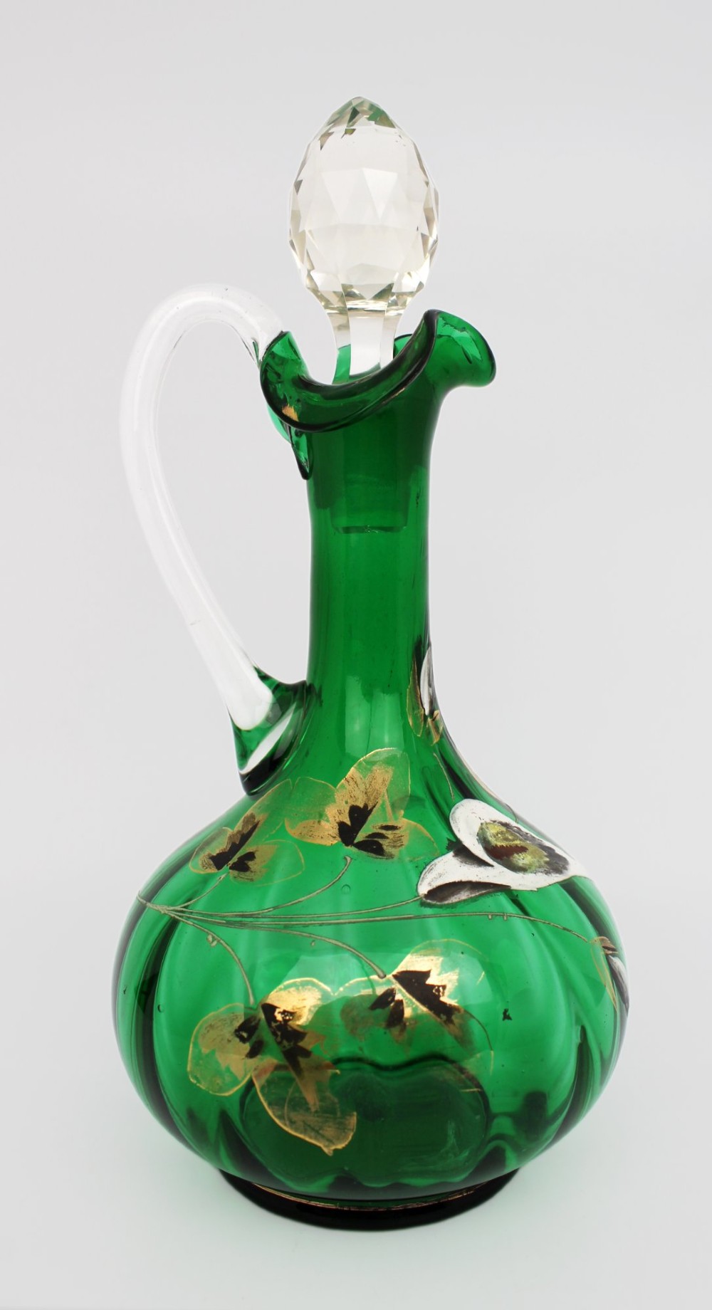 An emerald green glass claret jug featuring a ribbed bulbous body decorated with hand-painted gold floral motifs, a clear looped handle, and a faceted clear glass stopper.