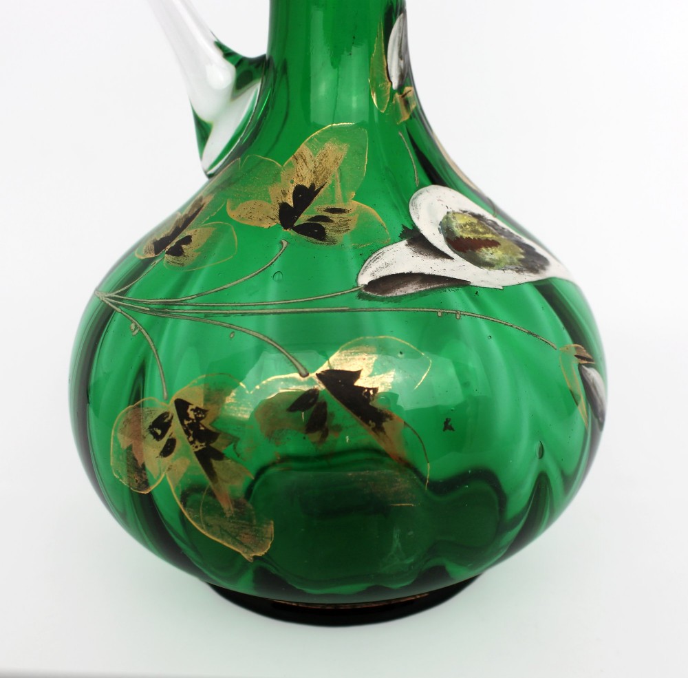 An emerald green glass claret jug featuring a ribbed bulbous body decorated with hand-painted gold floral motifs, a clear looped handle, and a faceted clear glass stopper. View IV