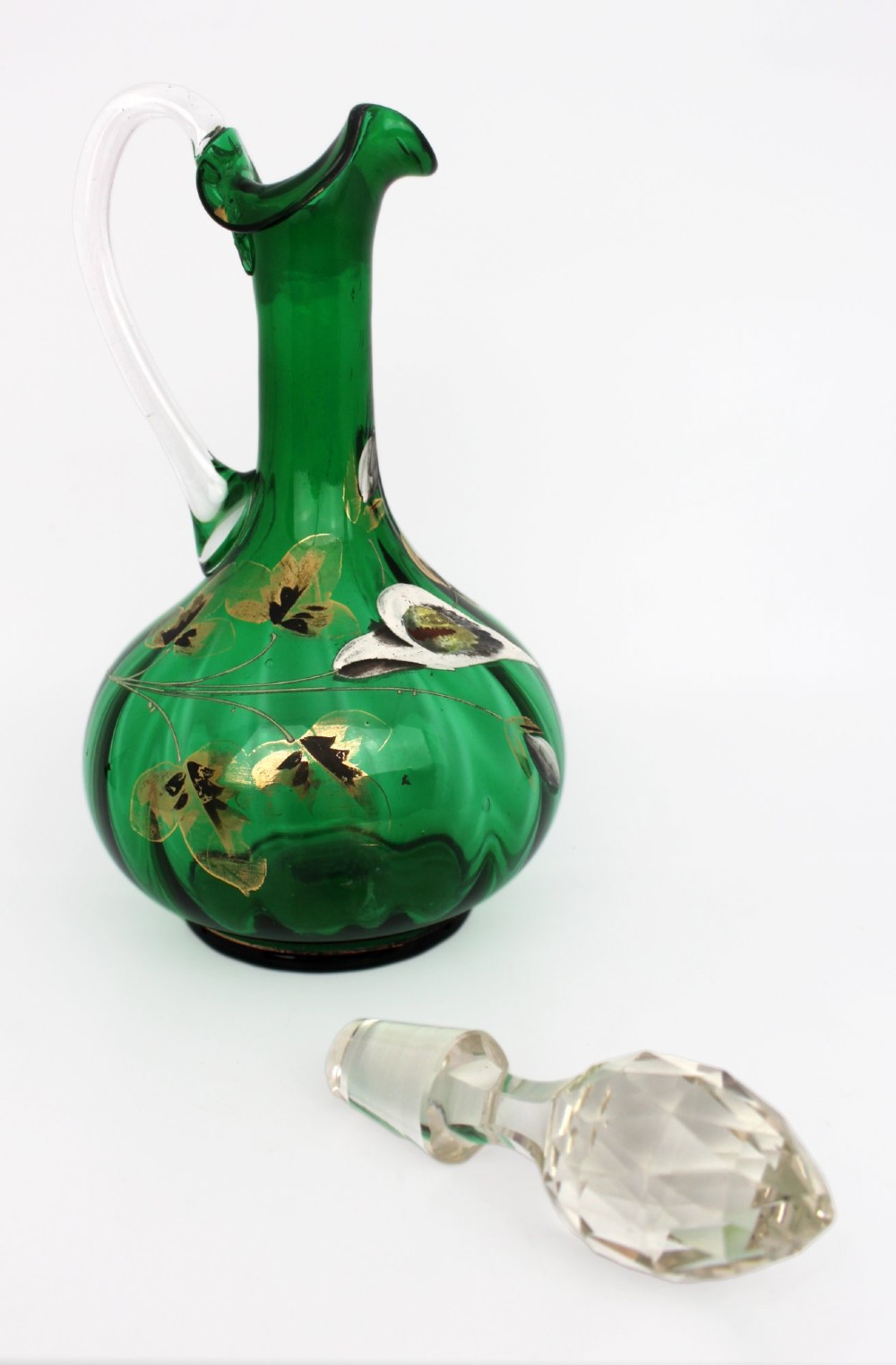 An emerald green glass claret jug featuring a ribbed bulbous body decorated with hand-painted gold floral motifs, a clear looped handle, and a faceted clear glass stopper. View V