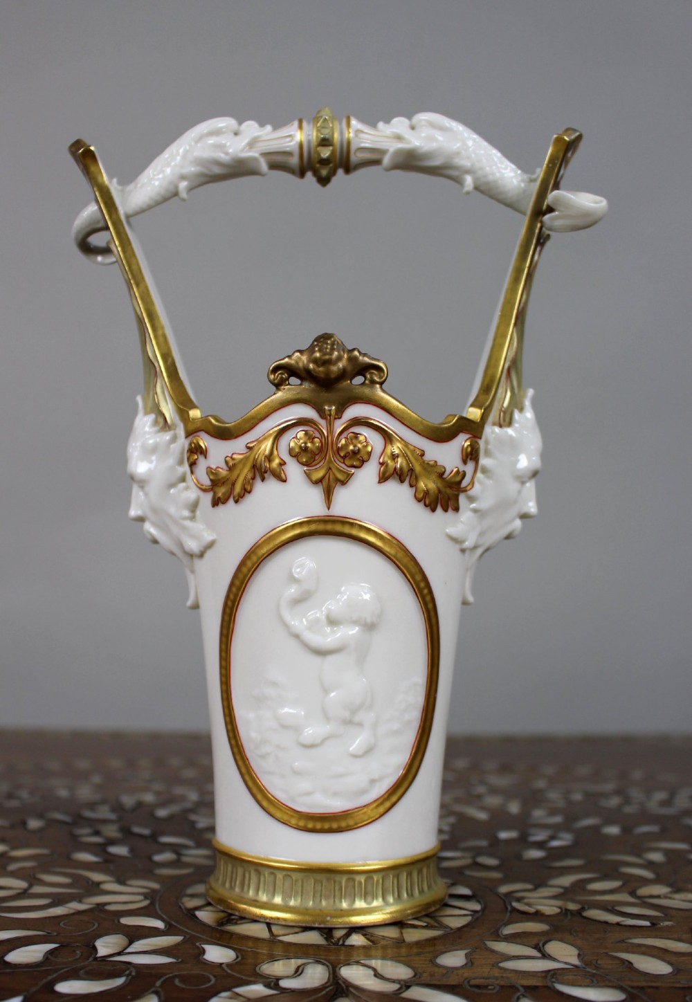 A white porcelain vase featuring a central white relief medallion of a cherub, gold gilded borders, raised side mask faces, and an overhead dolphin handle.