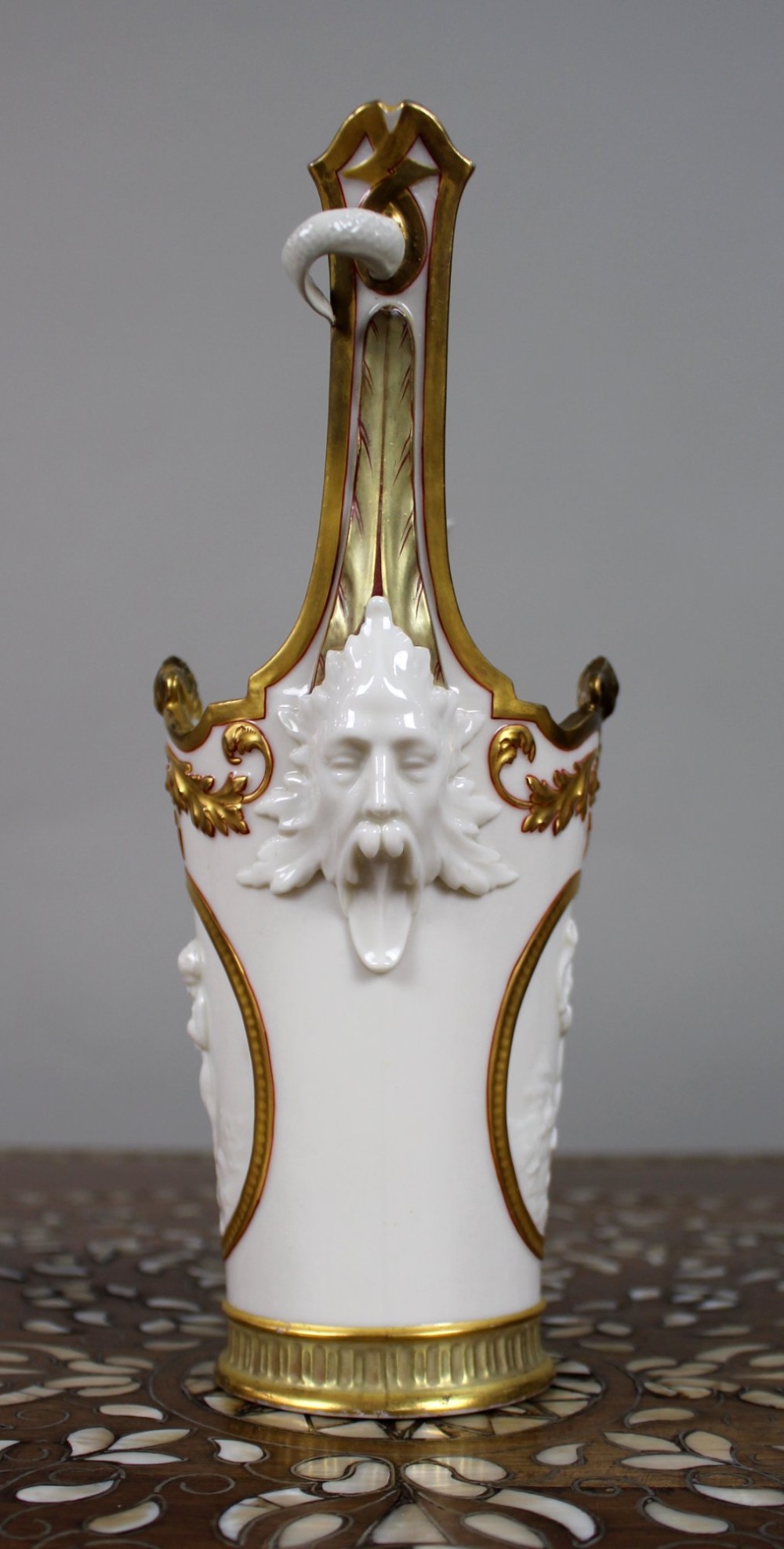 A white porcelain vase featuring a central white relief medallion of a cherub, gold gilded borders, raised side mask faces, and an overhead dolphin handle. View II