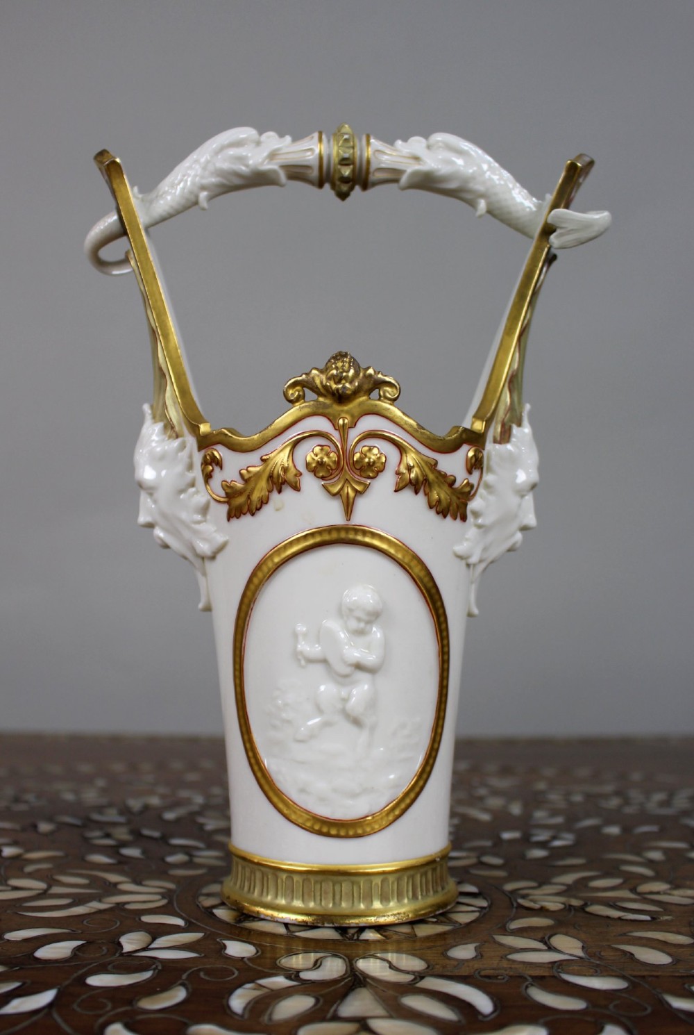 A white porcelain vase featuring a central white relief medallion of a cherub, gold gilded borders, raised side mask faces, and an overhead dolphin handle. View III