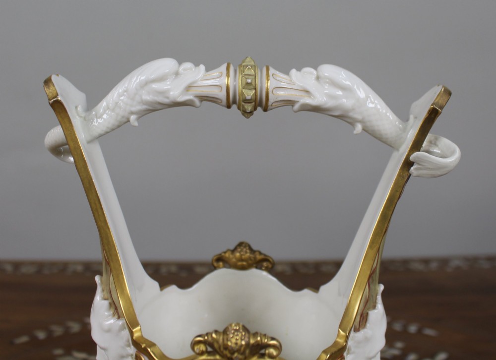 A white porcelain vase featuring a central white relief medallion of a cherub, gold gilded borders, raised side mask faces, and an overhead dolphin handle. View V