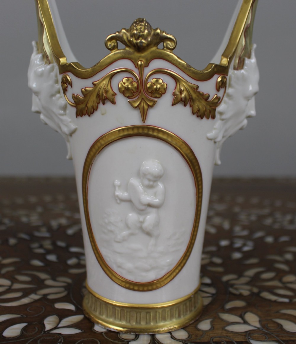 A white porcelain vase featuring a central white relief medallion of a cherub, gold gilded borders, raised side mask faces, and an overhead dolphin handle. View VI
