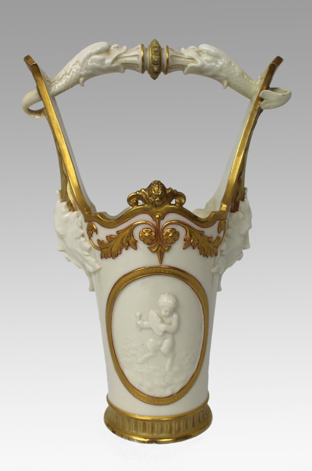 A white porcelain vase featuring a central white relief medallion of a cherub, gold gilded borders, raised side mask faces, and an overhead dolphin handle. View VIII