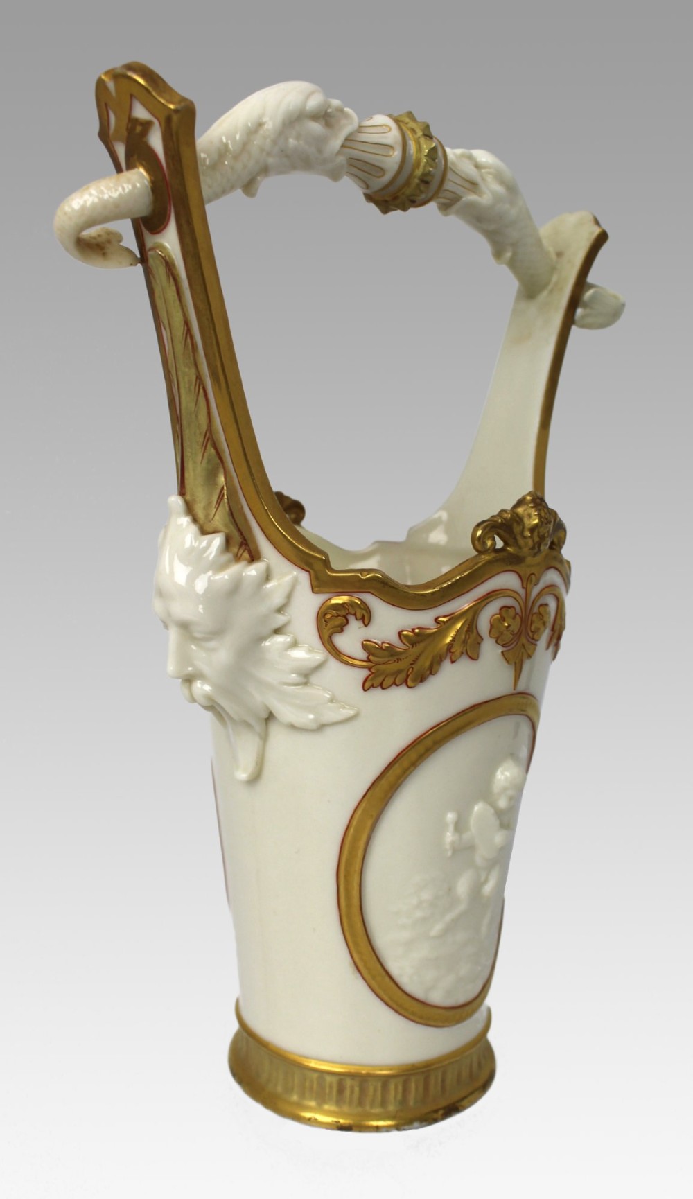 A white porcelain vase featuring a central white relief medallion of a cherub, gold gilded borders, raised side mask faces, and an overhead dolphin handle. View IX