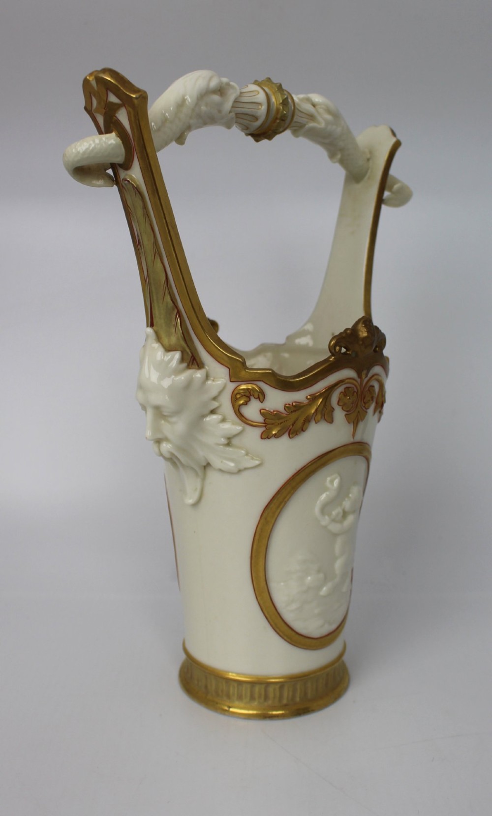 A white porcelain vase featuring a central white relief medallion of a cherub, gold gilded borders, raised side mask faces, and an overhead dolphin handle. View XII
