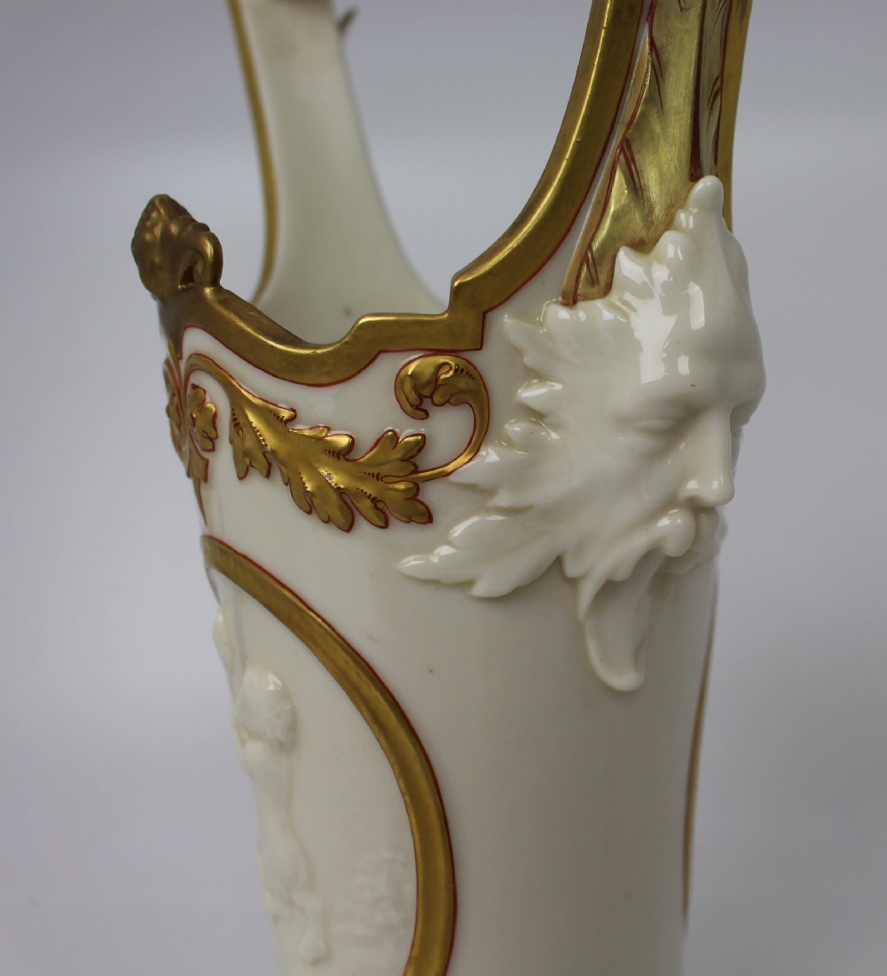 A white porcelain vase featuring a central white relief medallion of a cherub, gold gilded borders, raised side mask faces, and an overhead dolphin handle. View XIV