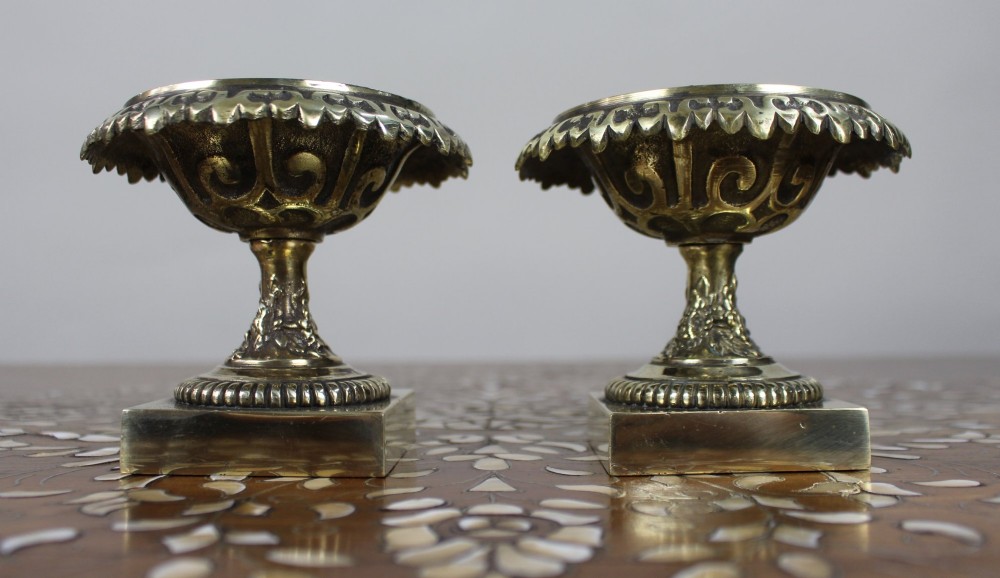 A pair of small antique bronze and brass urns featuring scalloped rims, relief scrollwork, and flared pedestals on square bases.