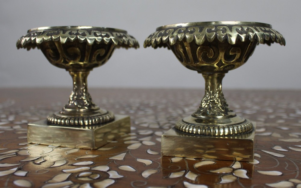 A pair of small antique bronze and brass urns featuring scalloped rims, relief scrollwork, and flared pedestals on square bases. View V