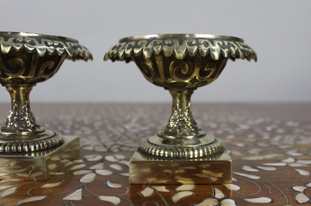 A pair of small antique bronze and brass urns featuring scalloped rims, relief scrollwork, and flared pedestals on square bases. View VII