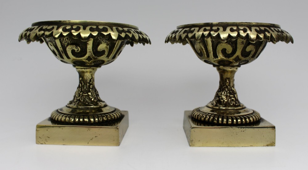 A pair of small antique bronze and brass urns featuring scalloped rims, relief scrollwork, and flared pedestals on square bases. View VIII