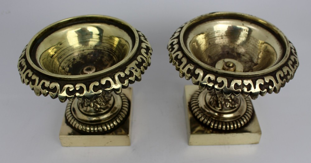 A pair of small antique bronze and brass urns featuring scalloped rims, relief scrollwork, and flared pedestals on square bases. View IX