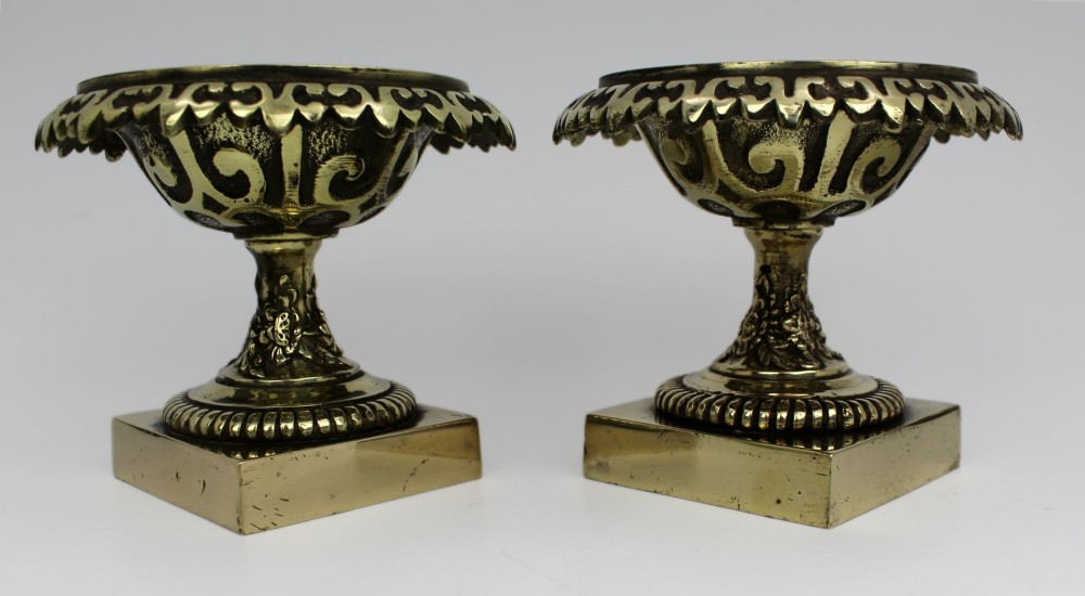 A pair of small antique bronze and brass urns featuring scalloped rims, relief scrollwork, and flared pedestals on square bases. View X