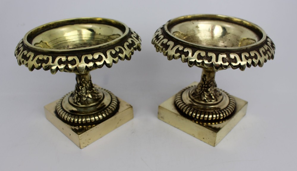 A pair of small antique bronze and brass urns featuring scalloped rims, relief scrollwork, and flared pedestals on square bases. View XI
