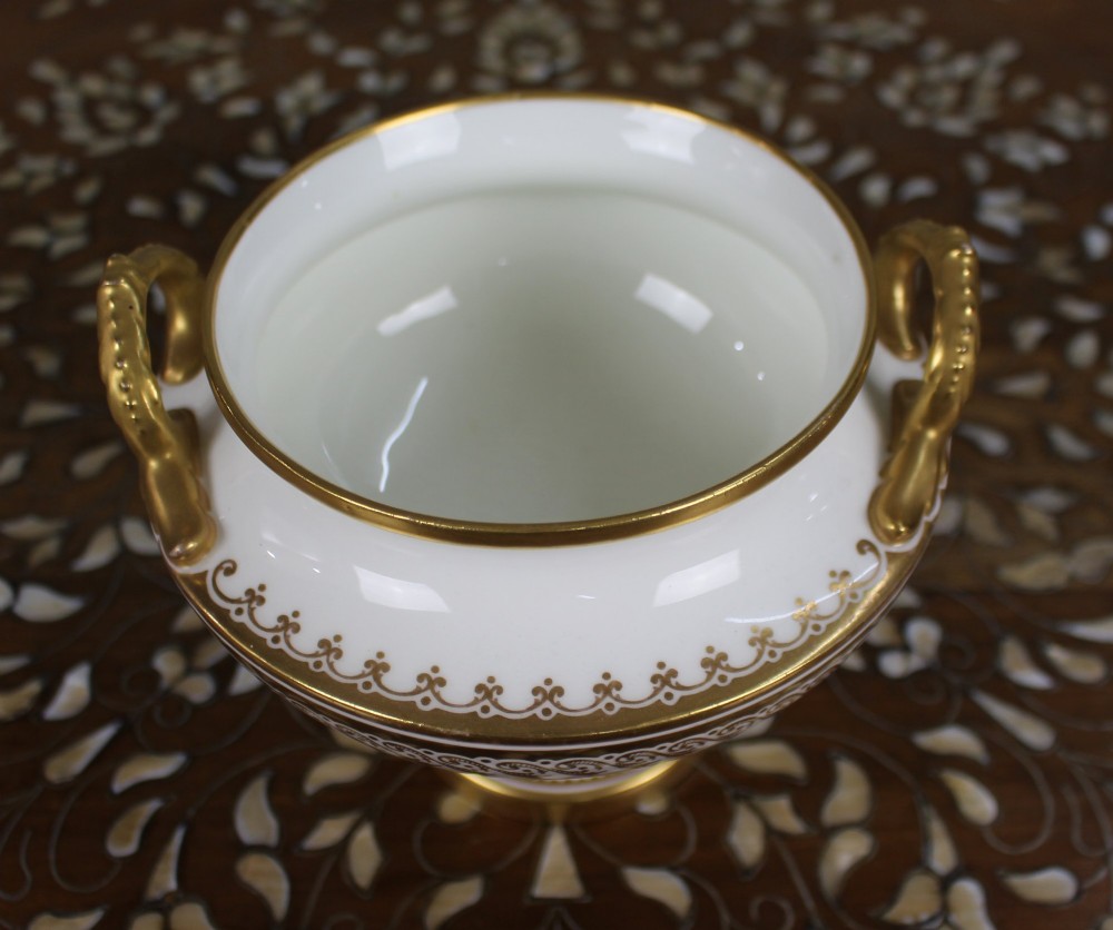 A 19th-century white porcelain urn featuring gilded side handles, a classical scroll frieze, and a fluted pedestal foot. View II