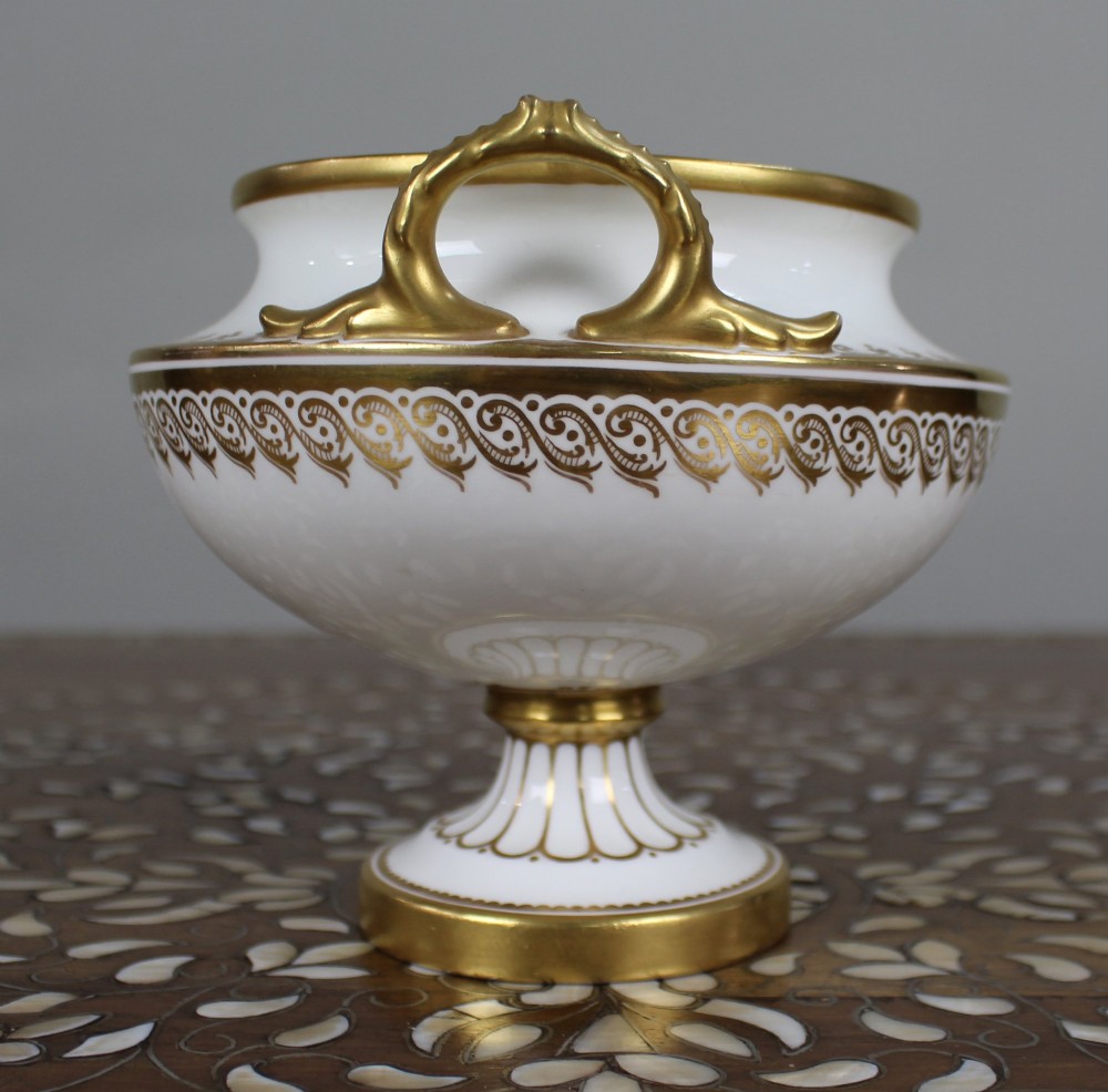 A 19th-century white porcelain urn featuring gilded side handles, a classical scroll frieze, and a fluted pedestal foot. View VI