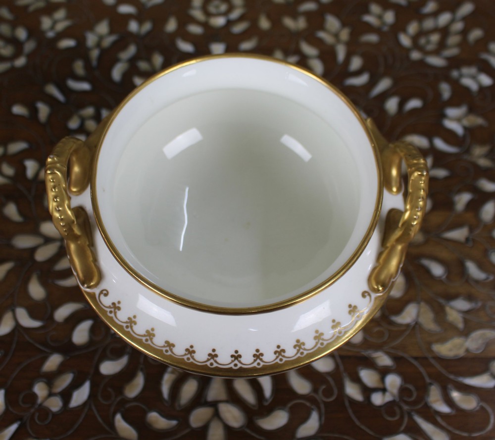 A 19th-century white porcelain urn featuring gilded side handles, a classical scroll frieze, and a fluted pedestal foot. View IX