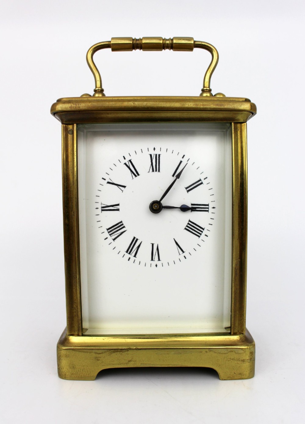 An antique brass carriage clock featuring rectangular glass side panels, a white enamel dial with black Roman numerals, and a folding top handle.