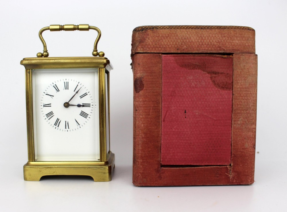 An antique brass carriage clock featuring rectangular glass side panels, a white enamel dial with black Roman numerals, and a folding top handle. View IX