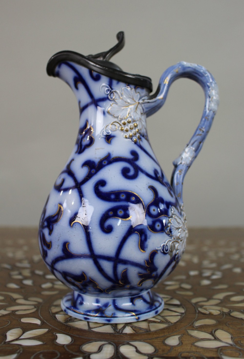 A Victorian relief-moulded flow blue earthenware jug decorated with cobalt scrollwork, white floral details with gold accents, and a hinged pewter lid.