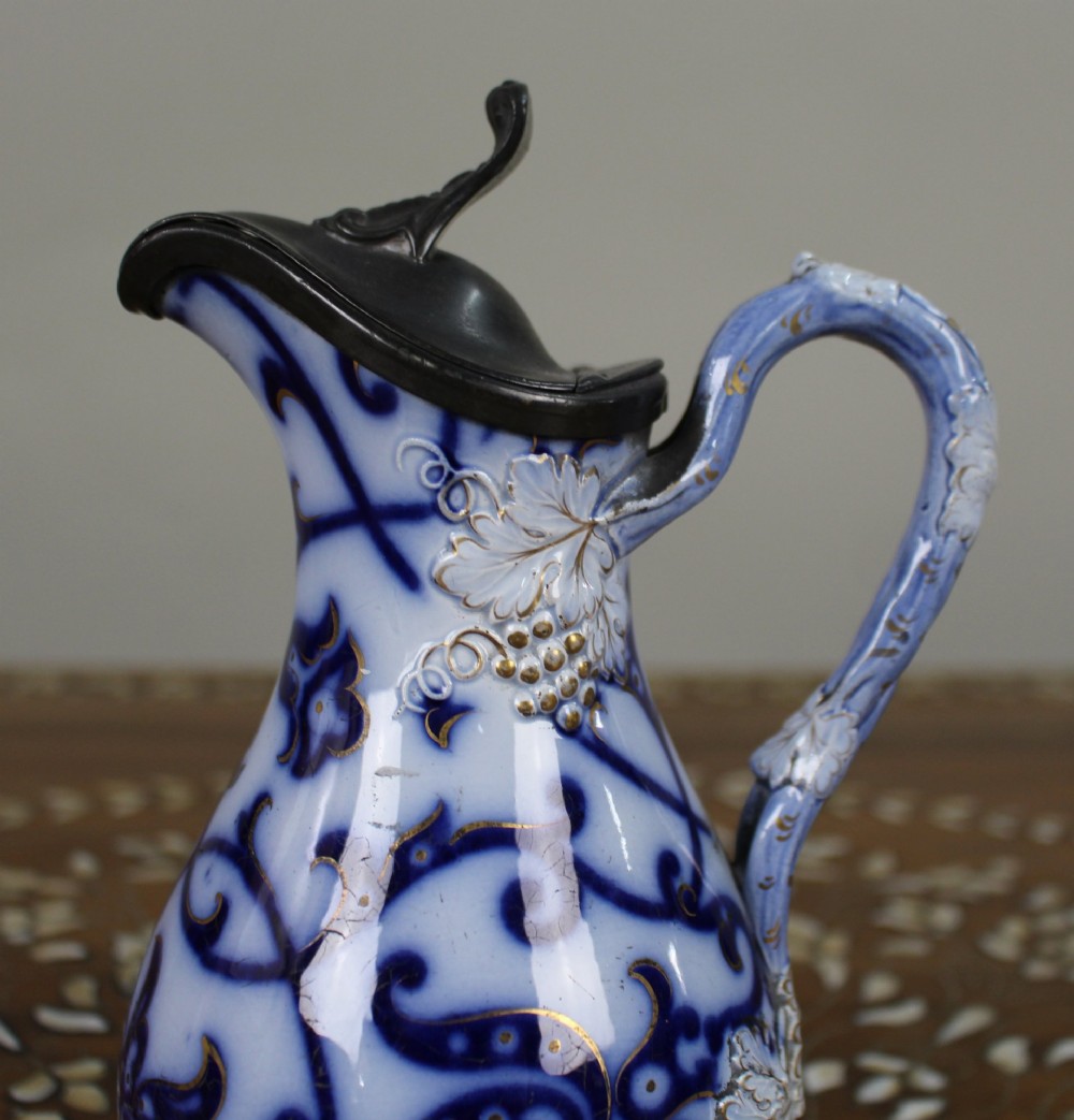 A Victorian relief-moulded flow blue earthenware jug decorated with cobalt scrollwork, white floral details with gold accents, and a hinged pewter lid. View II