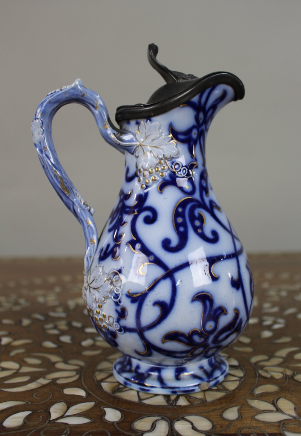 A Victorian relief-moulded flow blue earthenware jug decorated with cobalt scrollwork, white floral details with gold accents, and a hinged pewter lid. View V