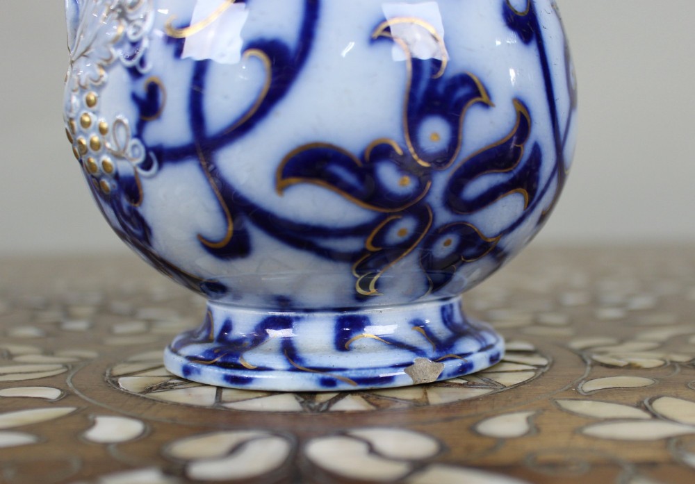 A Victorian relief-moulded flow blue earthenware jug decorated with cobalt scrollwork, white floral details with gold accents, and a hinged pewter lid. View VI