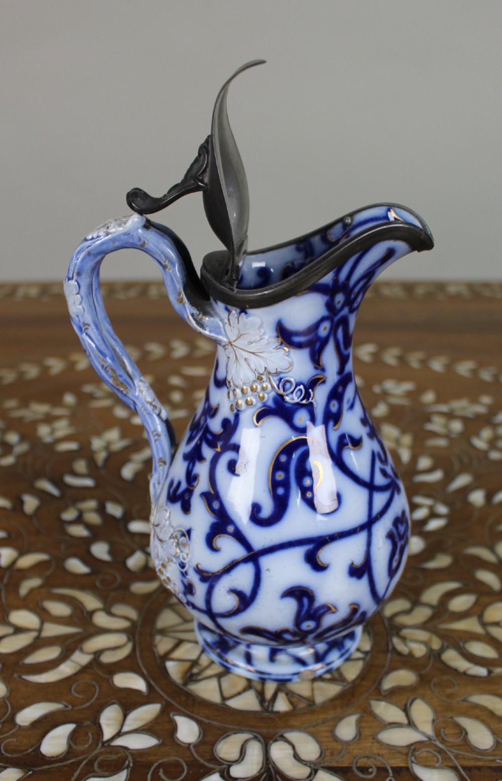 A Victorian relief-moulded flow blue earthenware jug decorated with cobalt scrollwork, white floral details with gold accents, and a hinged pewter lid. View VIII